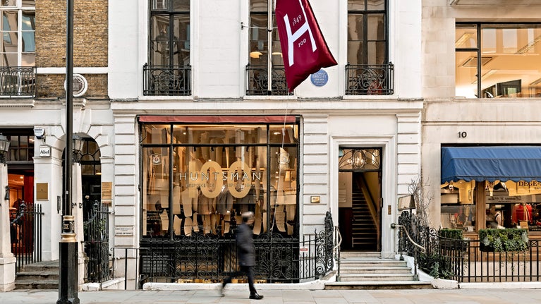 A view of Huntsman's shop front on Savile Row, their flag flying and a man is walking past on the pavement.